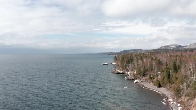 aerial drone view flying along shoreline of lake superior with forest and small cabin on hill in woods conifer forest in minnesota in winter
