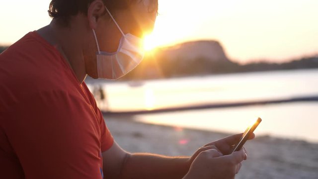COVID -19. Man wearing mask on sits on the beach uses mobile phone at sunset time. Coronavirus epidemic