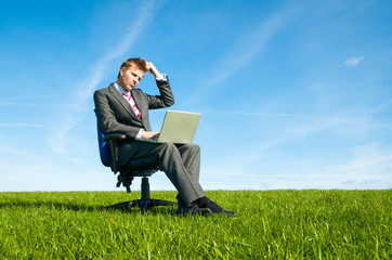 Thinking businessman sitting outdoors on an office chair working on a laptop scratching his head looking up in a sunny green meadow