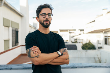 Confident stylish guy with tattoos posing on apartment balcony or terrace. Young man in glasses...