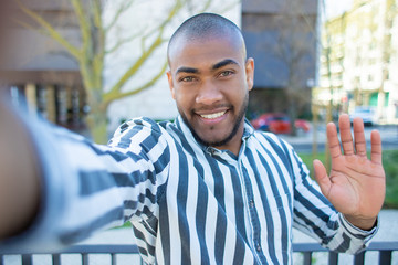 Handsome smiling African American man waving to camera. Cheerful young guy holding digital device in outstretched arm while posing for selfie. View from camera. Self portrait concept