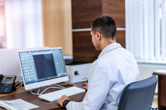 Doctor At His Desk With Laptop Computer. Modern Hospital Office. Photo From The Back. Results On Screen.