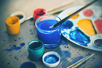 On a wooden table, there is a creative mess - in the mess are gouache paints, a palette and brushes.