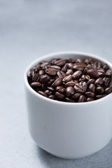A closeup view of a white mug full of coffee beans against a white marble background.