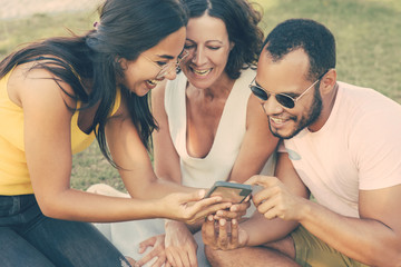 Cheerful multiethnic friends using smartphone. Smiling young man and women sitting together and using mobile phone in park. Technology concept