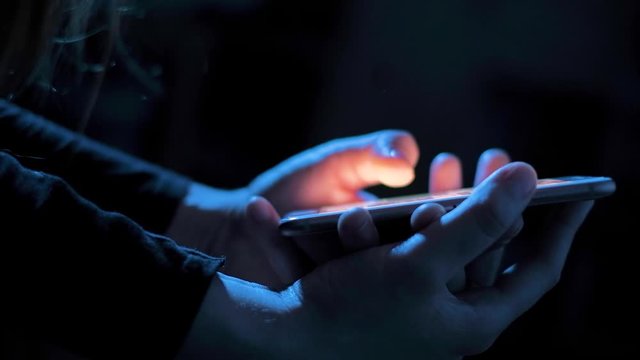 Young woman browsing internet on smart phone at night; girls finger tapping keypad, scrolling touchscreen of mobile device, blue low light; close up on hands side view