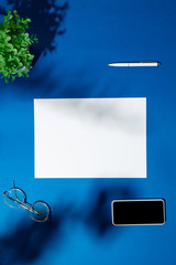 Sheets, gadgets and work tools on a blue table indoors. Creative, cozy workplace at home office, inspirational mock up with plant shadows on surface. Concept of remote office, freelance, atmosphere.