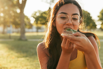 Excited Latin woman enjoying takeaway dinner outdoors. Beautiful young woman sitting on grass and eating pizza slice. Fast food concept