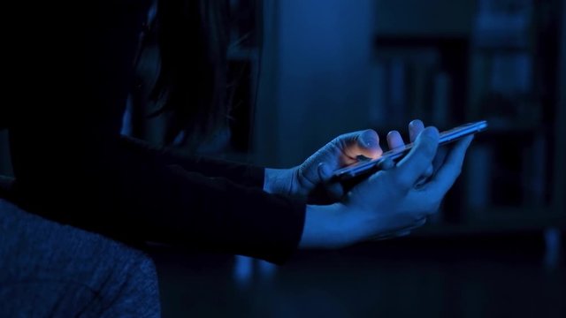 Side view close up of young woman browsing internet on smart phone at night; girls fingers tapping keypad, scrolling touchscreen of mobile device, blue low light; sleeplessness insomnia concept 