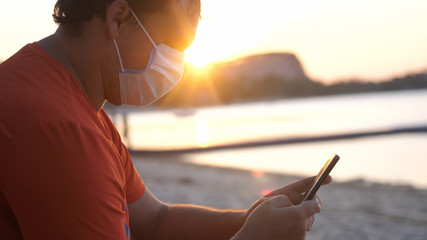COVID -19. Man wearing mask on sits on the beach uses mobile phone at sunset time. Coronavirus...