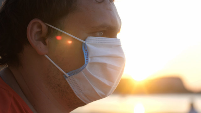 A masked young sad man sits on the beach at sunset time. Coronavirus epidemic