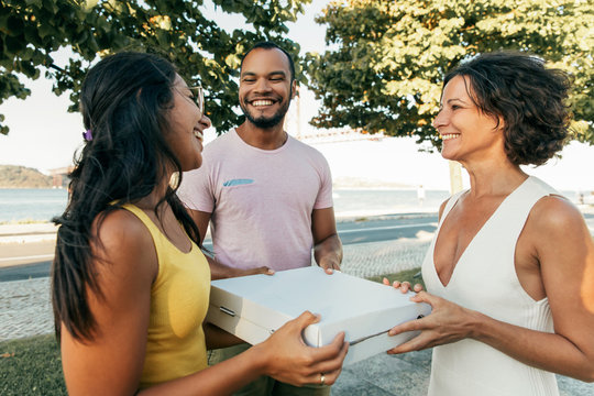 Joyful happy closed friends ready for takeaway dinner outdoors. Man and women standing in park, holding pizza boxes together, talking and laughing. Eating together concept