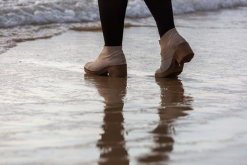 Girl's legs walking on the beach near water line in cream-colored winter shoes and black tights, water reflection 