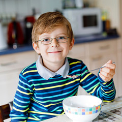 Adorable Happy little blond kid boy with glasses eating homemade cereals for breakfast or lunch....
