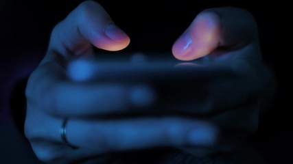 Young woman texting chatting on smart phone at night; girls finger tapping keypad, writing message on mobile devices touchscreen, blue low light; extreme close up top view