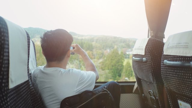 Back View Of Traveler Tourist Young Man Sitting On The Bus And Looking Outside Window To See Nature Landscape And Take A Photo With Light Effect Background. Tourist Travel Journey Adventure Concept