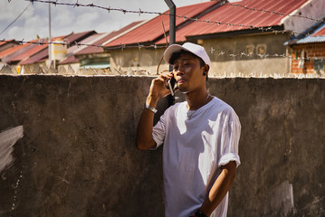 A young, brown-skinned Indonesian man dressed entirely in white with a cap, watch, and bracelets stands next to a grey concrete wall with glass fences talking to his smartphone. Face profile side.Asia