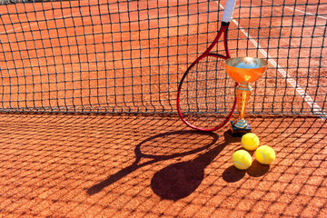 Trophy, professional tennis racket and balls on a red clay tennis court and net shadow. Champion or...