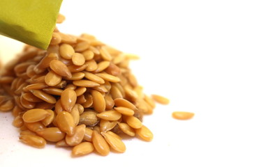 A close up photograph of golden linseeds, flax seeds, in a pile, being poured from a packet, isolated against a white background