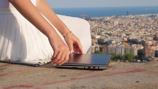 Girl press button on keyboard then turn off laptop by folding down screen, closeup shot of notebook. Young woman sitting at parapet on top point, blurred Barcelona cityscape on background