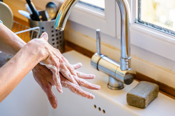 Close-up view on the hands of a woman washing her hands thoroughly with soap under the faucet of the kitchen sink.