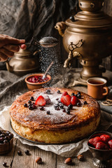 Woman hand sprinkles icing sugar on a cheesecake with berries on rustic wooden table with samovar and teapot, complex composition. Sweet dessert, close up