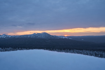 Winter forest with snow-covered fir trees high in the mountains. Dawn with bright colors on the horizon far away in the mountains. Golden clouds with the first rays of the sun.