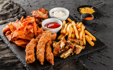 French fries potatoes, grilled meat and fish, chicken nuggets, spicy potato chips, ketchup and mayonnaise on black stone background. Hot fast food snacks, close up