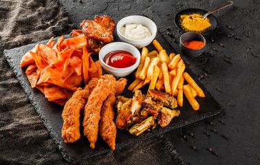 French fries potatoes, grilled meat and fish, chicken nuggets, spicy potato chips, ketchup and mayonnaise on black stone background. Hot fast food snacks, close up
