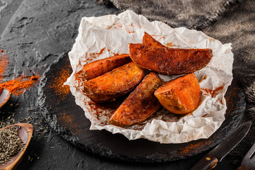 Fries potato wedges with red paprika and spice on paper over black stone background. Hot fast food, close up