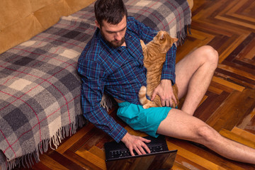 A bearded man is working on a laptop while sitting on the floor near the sofa.