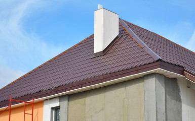 Close-up on a dark red metal tiled bonnet roof with a white chimney and stucco finish walls with scaffolding.