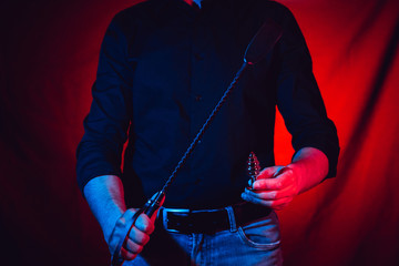 a man stands with a whip and an anal plug in his hands on a red background - obrazy, fototapety, plakaty