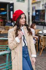 Obraz premium Beautiful young woman wearing red beret at city street of European city. Happy tourist girl walking outdoors. Spring portrait of pretty brunette female posing in old town.