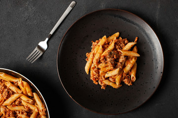 Fresh italian penne pasta bolognese with fork on dark plate and dark concrete background top view. 