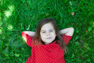 Top view of beautiful little girl is lying on a green grass, smiling and looking at the camera