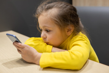 Naklejka premium little girl plays with the phone in a cafe