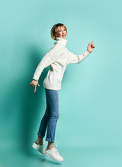 Blonde woman in white sweater, jeans and sneakers. She is laughing, jumping while posing sideways against blue studio background