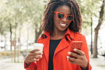 Smiling woman in sunglasses using smartphone. Content young African American woman holding coffee to go and using cell phone in park. Technology concept