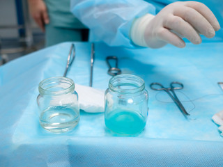 Sterile surgical instruments and glass containers with solutions on the table during a surgical operation. Above the table are the hands of a surgeon in latex gloves.