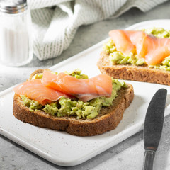 Bruschetta with avocado and salmon on a white ceramic plate on a light gray kitchen table. Toasted bread with avocado and red fish. The concept is helpful and a delicious Breakfast