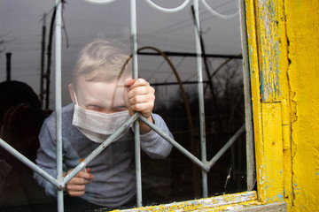 Boy kid blond European sitting in a medical mask in front of the window of the house. The child is angry that he can't go out with friends during the pandemic. 