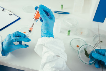 cropped view of biochemist taking medicine with syringe near colleague holding petri dish with green leaf