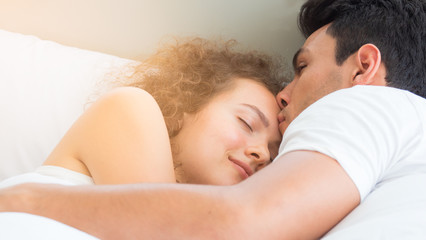 Fototapeta na wymiar Young couple, lovers or newly weds sleeping on white bed in the morning. Man give morning kiss to woman. Love and bonding