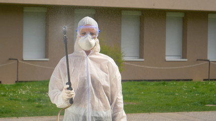 PORTRAIT: Woman in a white biohazard suit sprays disinfectant at the camera.