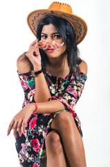 Young woman in a floral pattern dress wearing a cowboy hat and sitting cross legged with a slight emphasis to her right and perching a colorful spectacle on her nose with a stylish attitude 