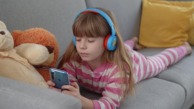 Cute little girl with headphones plays on a smartphone on a sofa