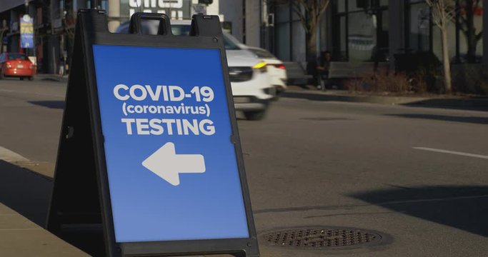 A sign on a sidewalk of a large city shows pedestrians and passing cars where the local COVID-19 testing center is.	