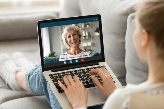 Back view of young woman sit relax on couch at home talk on video call with smiling mature mother, millennial girl rest have pleasant conversation with happy senior mom, use webcam on computer