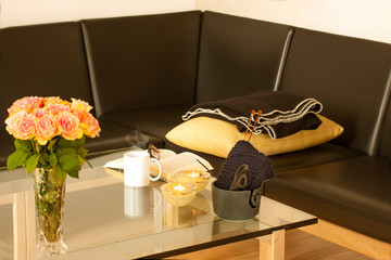 Book, glasses, roses, knitware and a mug with tea on modern glass table. Living room interior home decor concept with leather sofa.